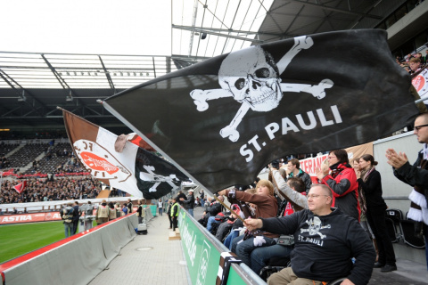 Fans im Rollstuhl auf ihren Plätzen direkt am Spielfeldrand schwenken große Fahnen mit dem Totenkopf-Symbol und dem Vereins-Wappen.